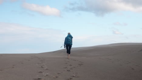 View-From-Behind-of-a-Blonde-Female-Hiker-with-Heavy-Backpack-Walking-Up-Barefoot-a-Sand-Dune-in-the-Desert-on-a-Beautiful-Summer-Day,-Råbjerg-Mile,-Denmark