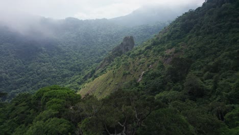 Cloudy-mountain-jungle-aerial-reveals-rock-spire-on-steep-green-slope
