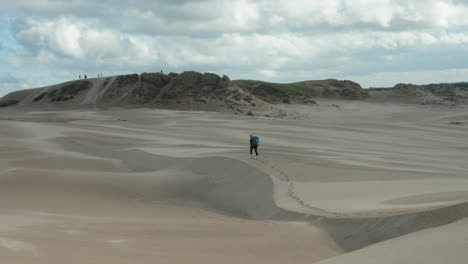 Beautiful-Wide-Shot-of-a-Lonely-Blonde-Female-Hiker-with-Heavy-Backpack-Walking-Up-Barefoot-a-Sand-Dune-in-the-Desert-on-a-Cloudy-Summer-Day,-Råbjerg-Mile,-Denmark