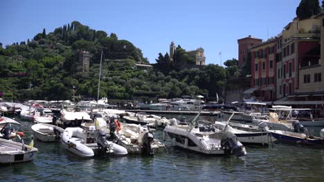 Boats-mooring-in-the-small-harbour-of-Portofino,-Italy-and-the-natural-olive-background-view-of-the-iconic-landmarks-of-Chiesa-di-San-Giorgio