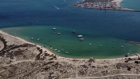 Bahía-De-La-Isla-Desierta,-Pequeños-Barcos-Amarrados-En-Aguas-Esmeralda-Con-La-Isla-Del-Faro-Al-Fondo,-Algarve