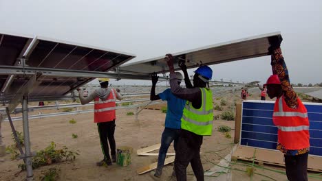Hombres-Colocando-Cuidadosamente-Paneles-Solares-Fotovoltaicos-Bifaciales-Sobre-Marcos-De-Acero-En-El-Proyecto-De-Granja-Solar-En-Gambia,-África-Occidental