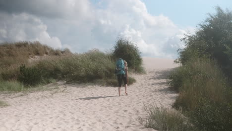 Wide-Shot-of-a-Blonde-Female-Hiker-with-Heavy-Backpack-Walking-Up-a-Sand-Dune-on-a-Beautiful-Summer-Day,-Råbjerg-Mile,-Denmark