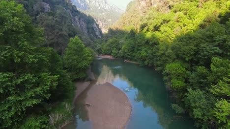 Aerial-fpv-drone-flies-through-river-at-base-of-canyon-as-explorer-dives-off-sand-bank-into-water