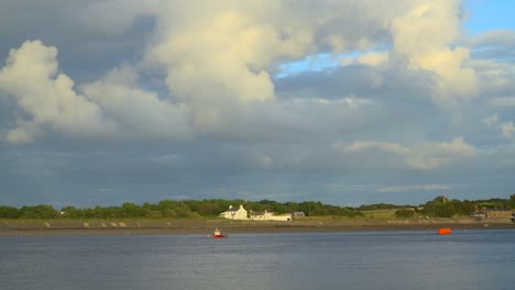 Stormy-sky-with-clouds-sweeping-by-as-tide-comes-in-on-foreground-river-with-moored-boat-and-riverside-cottage-at-sunset-on-summer-evening