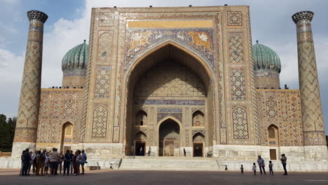 Exterior-of-Sherdor-Madrasah,-Landmark-of-Samarkand-Uzbekistan-and-People-in-Front-on-Sunny-Day