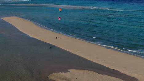 Playa-de-La-Barca,-Fuerteventura:-aerial-view-in-a-circle-over-the-beautiful-beach-where-people-practice-kitesurfing-on-a-sunny-day