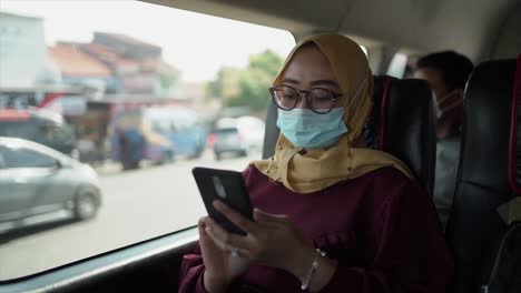 Positive,-Fröhliche-Junge-Frau-In-Freizeitkleidung-Sitzt-Auf-Dem-Mittelsitz-Des-Autos,-Trägt-Maske,-Brille,-Kopftuch-Und-Schaut-Weg,-Nachdem-Sie-Ihr-Mobiltelefon-Benutzt-Hat