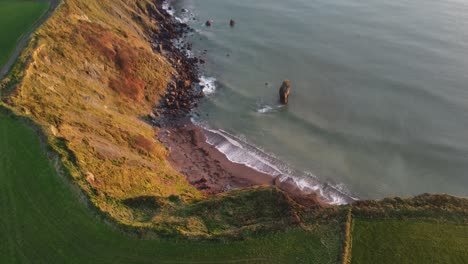 Drone-revealing-sea-cliffs-beach-and-sea-stack-at-The-Copper-Coast-Waterford-Ireland-at-golden-hour-on-a-winter-day