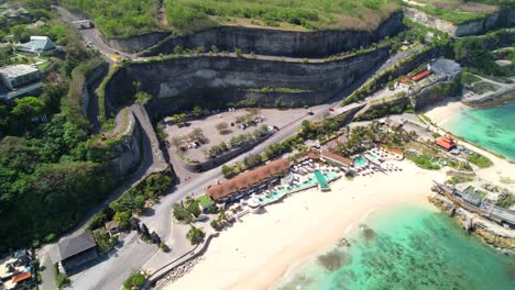 Aerial-Descending-Revealing-Exotic-Beach-Clubs-in-Melasti-Beach,-Winding-Steep-Cliff-Road-to-Pantai-Melasti-Ungasan-Resort-in-Uluwatu,-Bali