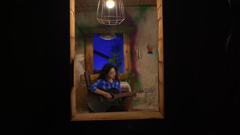 Junge-Frau-Spielt-Nachts-Gitarre,-Wie-Durch-Ein-Fenster-Sichtbar