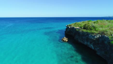 Drone-descends-along-rocky-shoreline-to-clear-blue-turquoise-waters-of-curacao