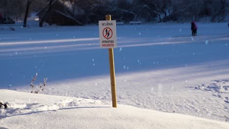 Danger-sign-forbids-to-step-on-lake-ice-but-people-still-walking-in-background,-static-view-during-snowfall
