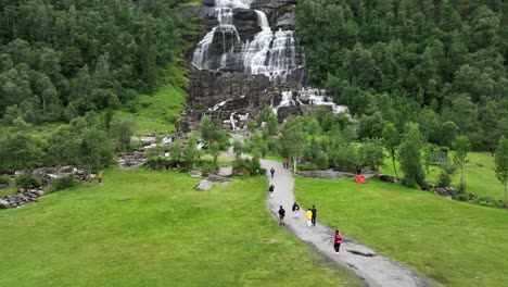 Turistas-Caminando-Hacia-La-Majestuosa-Cascada-Tvindefossen-En-Voss-Noruega---Vía-Aérea-Con-Inclinación-Lenta-Hacia-Arriba-Desde-La-Gente-En-El-Camino-Hasta-La-Vista-Completa-De-La-Cascada