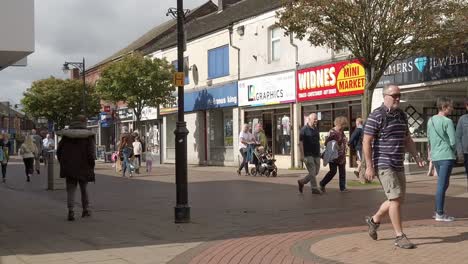 Slow-motion-people-walking-in-British-town-passing-general-shops-during-high-inflation-cost-of-living-crisis,-Panning-right