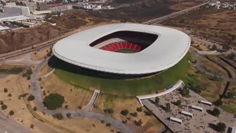 Aerial-tilt-up-shot-of-cars-at-parking-area-in-front-of-stunning-Akron-Stadium-during-sunny-day-in-Guadalajara,-Jalisco,-Mexico