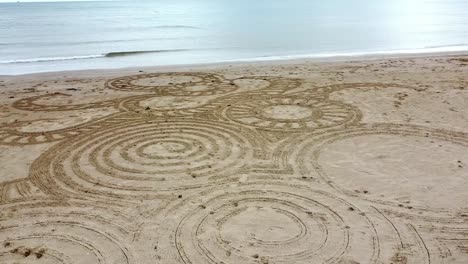 Luftaufnahme-Eines-Kreisenden-Mannes,-Der-Am-Windigen-Walisischen-Goldenen-Strand-Ein-Interessantes-Zen-Sand-Kunstdesign-Kreiert