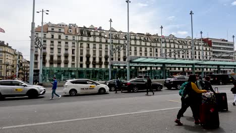 Menschen,-Die-Mit-Rollenden-Koffern-Zum-Eingang-Des-Genfer-Bahnhofs-Gehen,-Mit-Straßenbahnhaltestelle-Im-Hintergrund