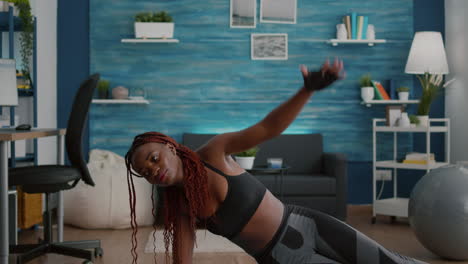 Flexible-Schwarze-Frau-Wärmt-Sich-Auf-Einer-Yoga-Karte-Im-Wohnzimmer-Auf,-Die-Auf-Der-Seitenplanke-Steht
