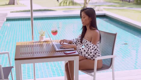 Thoughtful-ethnic-young-woman-using-laptop-at-poolside