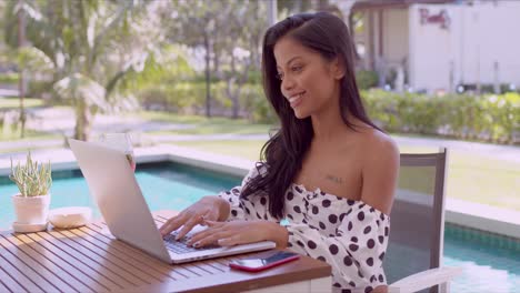 Happy-ethnic-young-woman-using-laptop-near-swimming-pool