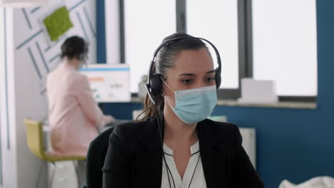 Close-up-of-executive-manager-with-face-mask-and-headset-working-on-laptop-computer
