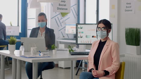 Portrait-of-manager-with-face-mask-sitting-on-chair-at-desk-table