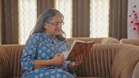 Happy-Indian-aged-woman-reading-book-and-drinking-tea