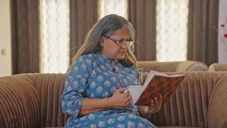 Sad-Indian-aged-woman-reading-book-and-drinking-tea