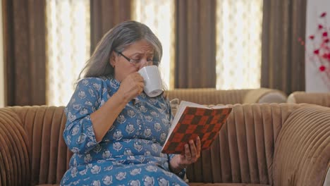 Angespannte-Indische-Frau-Im-Alter,-Die-Ein-Buch-Liest-Und-Tee-Trinkt
