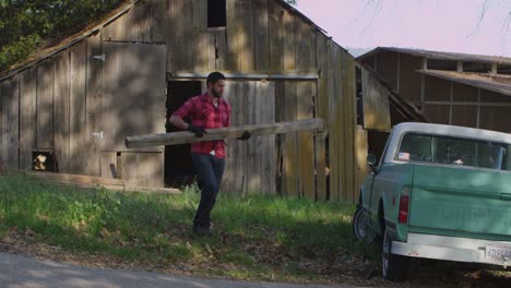 A-man-carries-a-board-to-a-pickup-truck-parked-outside-an-old-barn