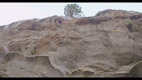 The-camera-tilts-up-from-a-climbing-partner-on-the-ground-to-reveal-a-rock-climber-high-on-a-rock-wall