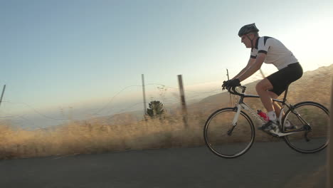 Traveling-shot-of-a-man-pedaling-a-mountain-bike-along-a-mountain-road-at-dusk