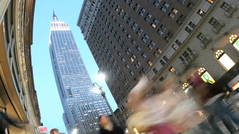 Das-Empire-State-Building-Wird-Von-Blauem-Himmel-Eingerahmt,-Da-Beschleunigter-Verkehr-Und-Fußgänger-Im-Vordergrund-Vorbeifahren