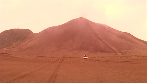 A-vehicle-motors-through-a-desert-landscape-as-a-volcanic-ash-cloud-forms-in-the-background-
