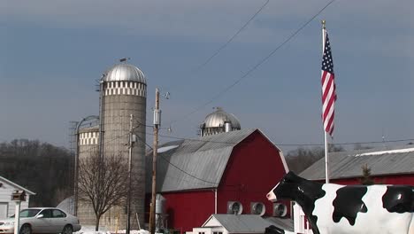 Die-Kamera-Erfasst-Eine-Amerikanische-Farm-Mit-Silos,-Rote-Scheune,-Amerikanische-Flagge-Und-Lebensgroße-Nachbildung-Einer-Kuh