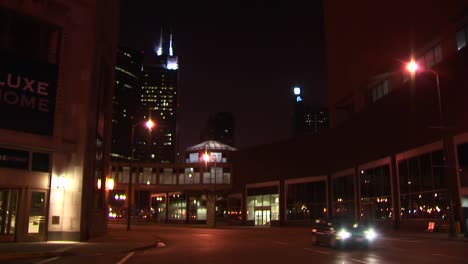 Nighttime-Urban-Scene-With-Taxis-And-Other-Vehicles-Traveling-Across-An-Intersection-With-Chicago-Landmark-The-Sears-Tower-In-The-Background