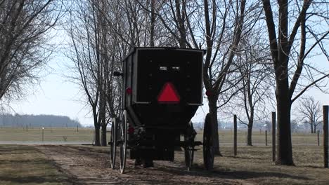 Ein-Amish-Pferd-Und-Ein-Buggy-Fahren-Mit-Ihren-Passagieren-Und-Einem-Warnenden-Roten-Dreieck-Auf-Der-Rückseite-Des-Buggys-In-Richtung-Hauptstraße-Road