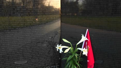 Una-Maceta-De-Lirios-Blancos-Sosteniendo-Una-Bandera-Roja-Se-Encuentra-Al-Pie-Del-Muro-Conmemorativo-De-Los-Veteranos-De-Vietnam-En-Washington-DC,-Cuya-Superficie-De-Granito-Refleja-El-Paisaje-Circundante