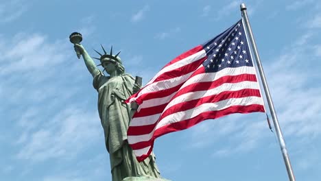 Die-Freiheitsstatue-Steht-Hoch-Vor-Blauem-Himmel,-Während-Eine-Amerikanische-Flagge-Im-Vordergrund-Weht