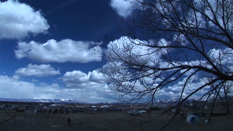 Weite-Aufnahme-Des-Himmels-In-Colorado-Mit-Autofriedhof-Und-Baumzweig-Im-Vordergrund-Und-Felsigen-Bergen-Im-Hintergrund