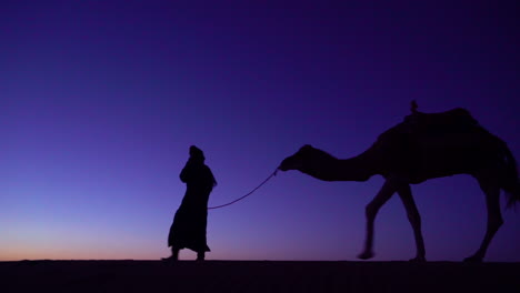 Eine-Klassische-Reiseabenteueraufnahme-Eines-Mannes,-Der-Ein-Kamel-In-Silhouette-über-Eine-Düne-Führt-Leading