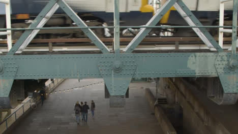 Fußgänger,-Die-Unter-Der-Brücke-Laufen,-Während-Der-Zug-Oben-Vorbeifährt
