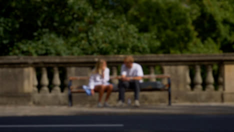 Defocused-Shot-of-People-and-Traffic-Moving-In-Front-of-Couple-Sat-On-Bench-01