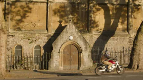 Wide-Shot-of-Motorcyclist-Riding-Past-Doors-to-St-Nicholas-Church-In-Bristol-