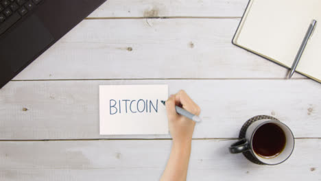 Top-Down-View-of-Woman-Writing-Bitcoin-On-Paper-with-Laptop-and-Notebook