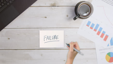 Top-Down-View-of-Woman-Writing-Failure-On-Paper-with-Business-Documents-and-Laptop