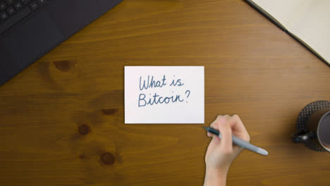 Top-Down-View-of-Woman-Writing-What-Is-Bitcoin-On-Paper-with-Notebook-and-Laptop