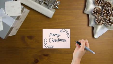 Draufsicht-Der-Hand,-Die-Frohe-Weihnachten-Auf-Papier-Mit-Weihnachtsgeschenken-Schreibt