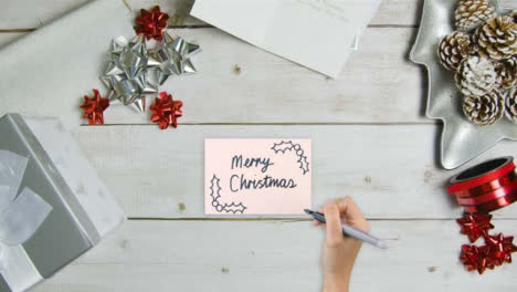 Draufsicht-Der-Hand,-Die-Frohe-Weihnachten-Auf-Papier-Mit-Weihnachtsdekorationen-Und--geschenken-Schreibt
